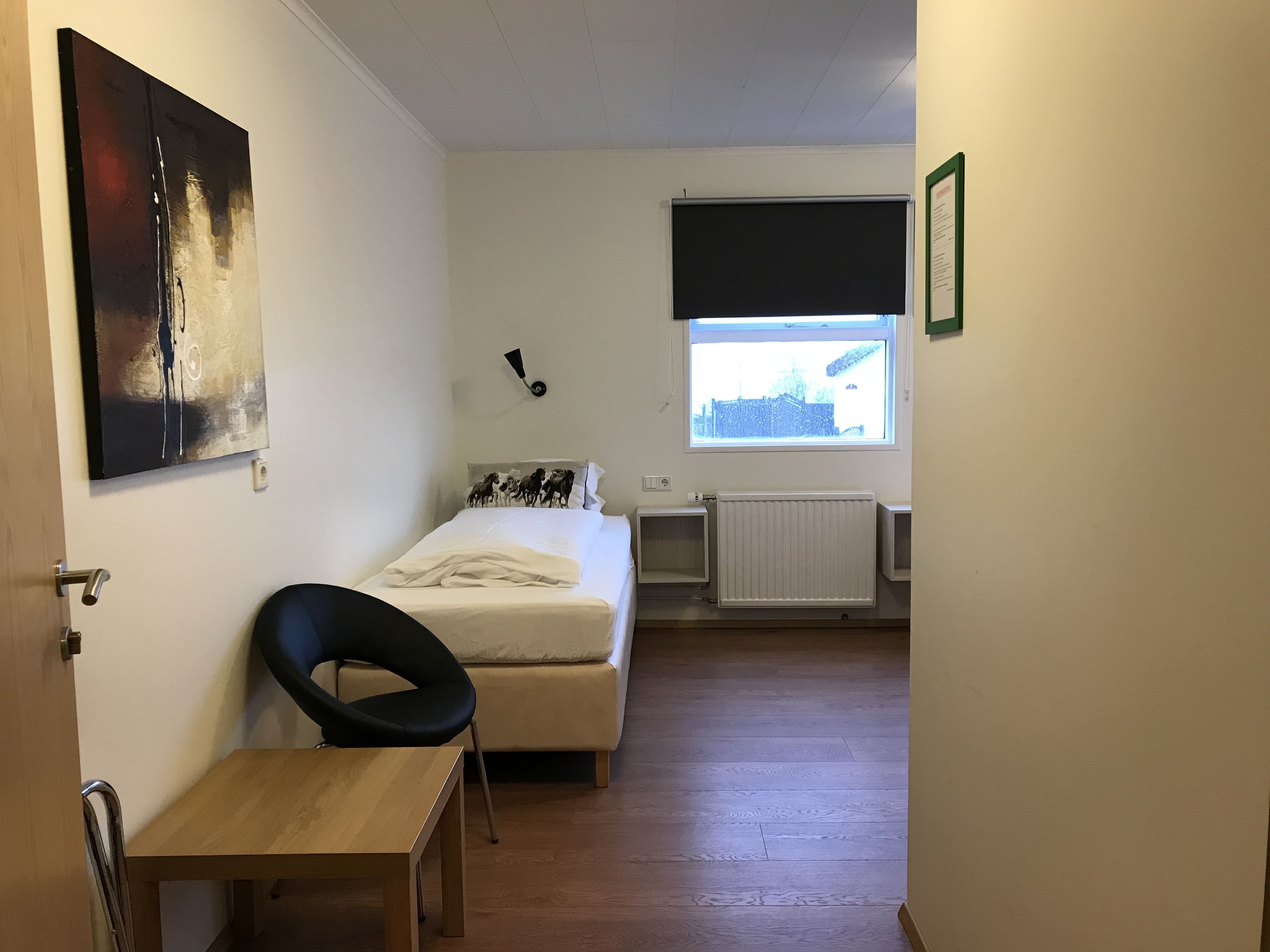 Une petite pièce bien éclairée, comprenant un lit, une chaise, une table, un radiateur et une fenêtre donnant sur les bâtiments environnants.