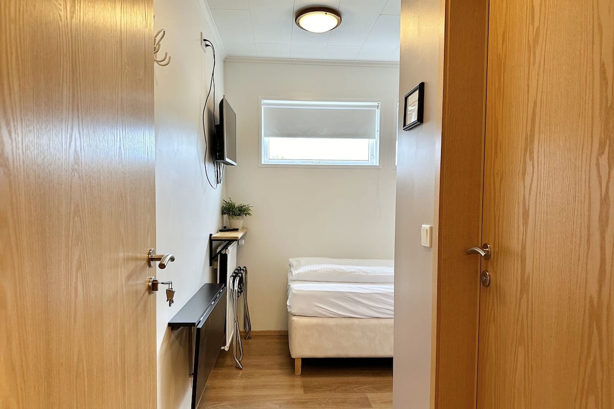 Ein schmaler Flur führt zu einem Schlafzimmer mit einem Bett, einem kleinen Tisch mit einer Pflanze, einem an der Wand montierten Fernseher und einem Fenster, das natürliches Licht hereinlässt.