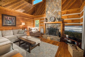 A log cabin interior with a stone fireplace, wooden walls, a sofa, a coffee table, and a television. - Peaceful Boone Log Cabin | Mountain Views, Fireplace, Loft & Pet Friendly (Boone)