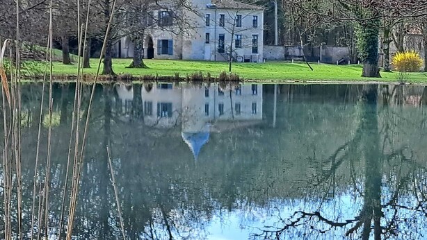 A serene lakeside view with a historic building featuring a blue dome, surrounded by trees and a calm water body reflecting the structure. - Lodge Hôtel Domaine De Sommedieue (Sommedieue)