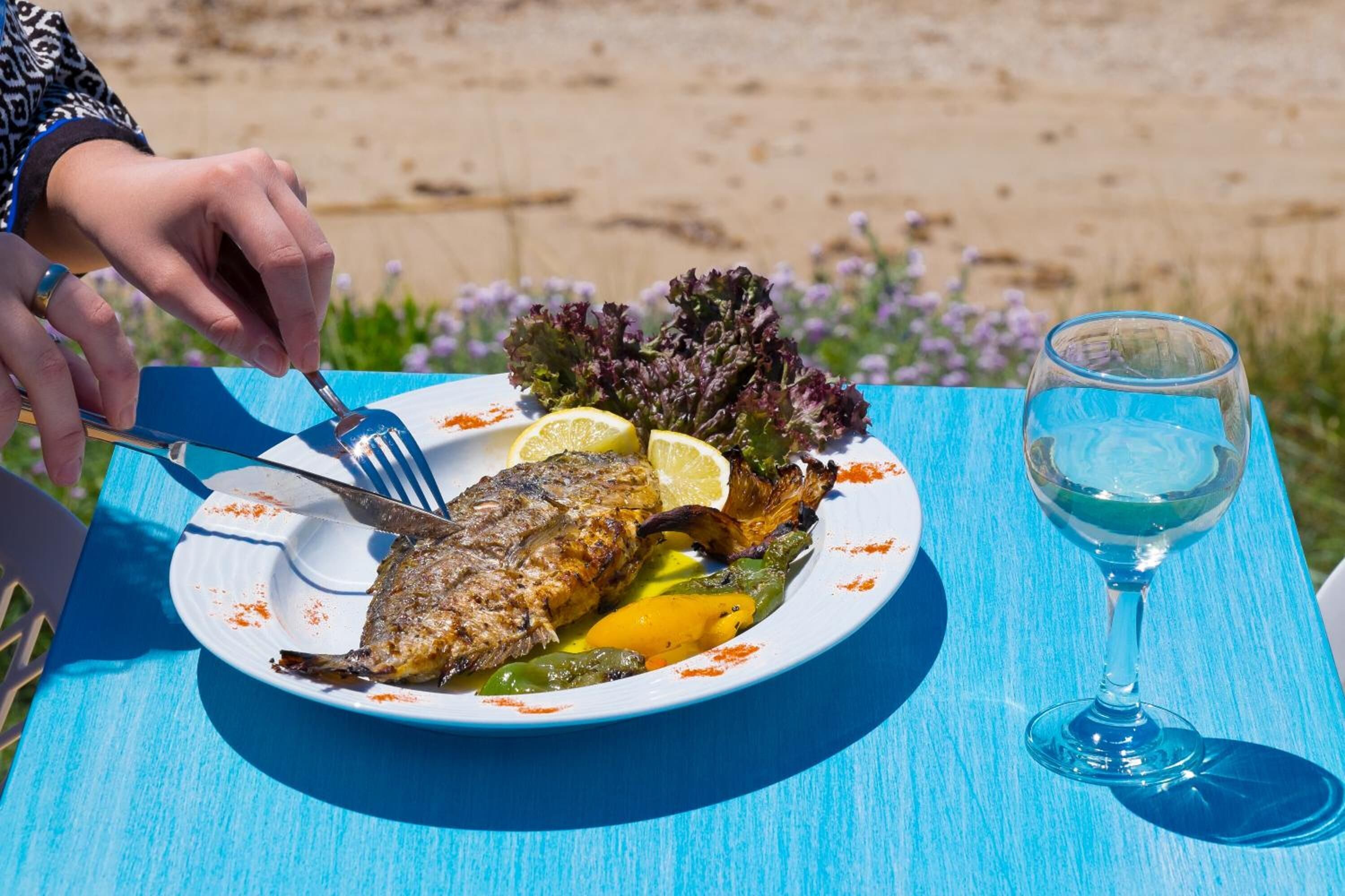 Un plato de pescado a la parrilla con verduras y rodajas de limón, acompañado de una copa de vino blanco, sobre una mesa azul al aire libre.