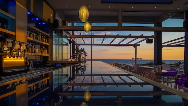 Un área de bar moderna con barandilla de vidrio que da a la vista del mar, con mostrador y estantes llenos de botellas.
