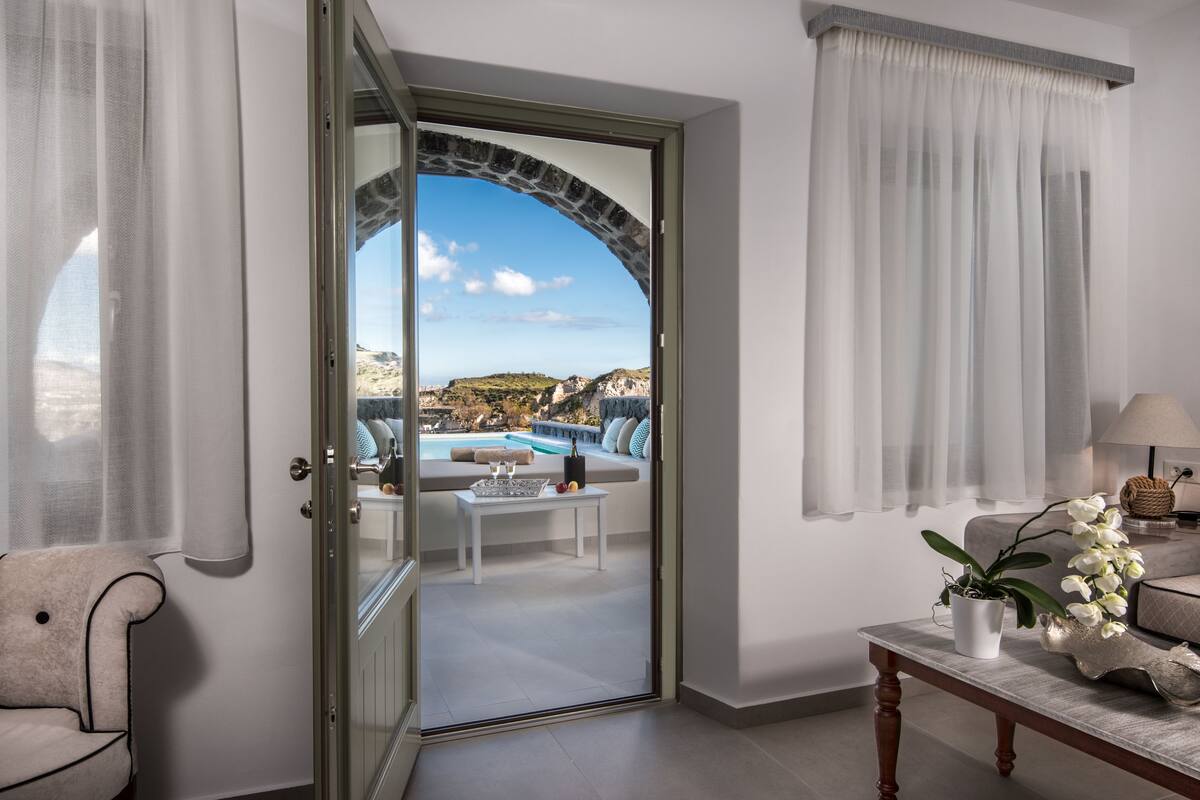 A room with a glass door leading to a balcony with a pool and a scenic view of cliffs.