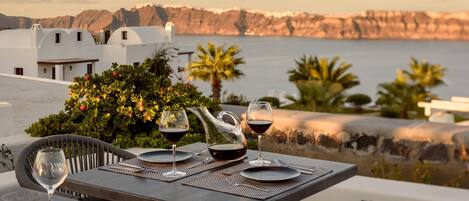 Mesa para dos con copas de vino y una jarra, con vista a un paisaje costero con edificios y acantilados.