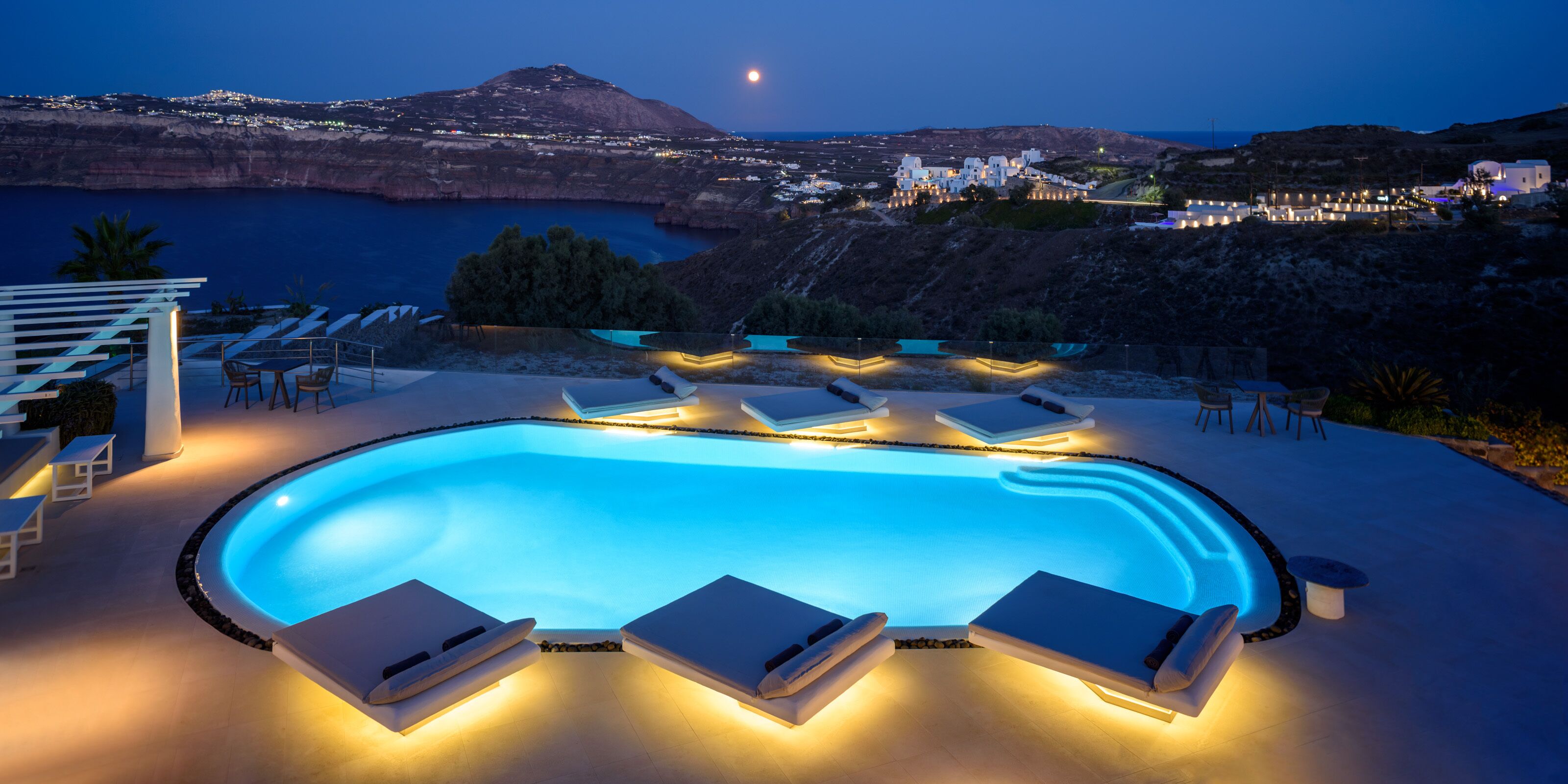 A swimming pool with lounge chairs, a view of the sea, and a mountainous landscape at dusk.
