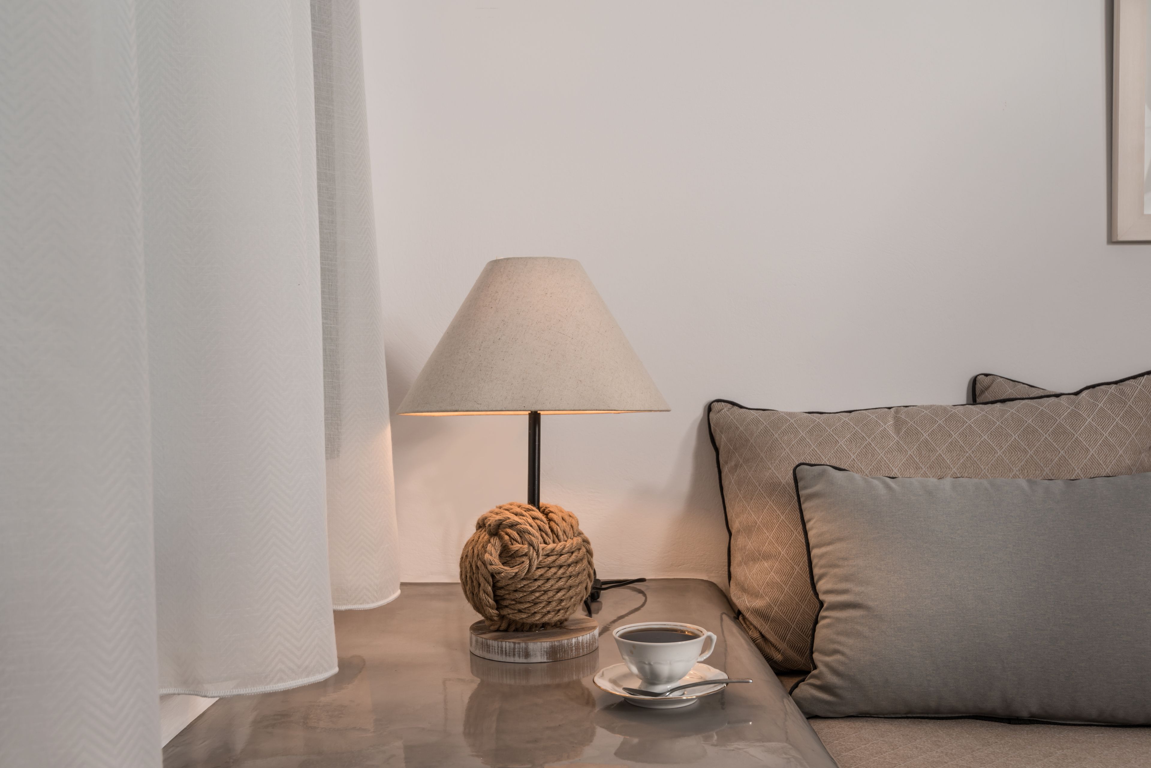 A modern room with a round, rope-wrapped lamp on a wooden coaster, a white cup on a saucer, and a sofa with patterned cushions.