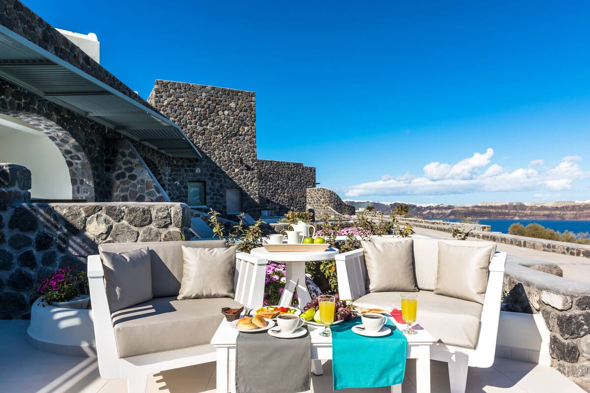 A sunlit outdoor seating area with a stone wall, a white cushioned bench, and a table set with breakfast items and a glass of orange juice.