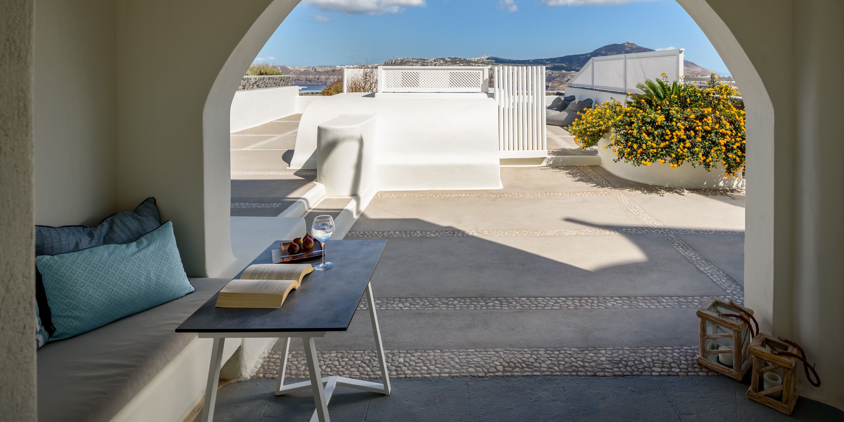 A cozy reading nook with a table, books, and a glass of water, overlooking a scenic view with a white fence and a yellow flower bush.