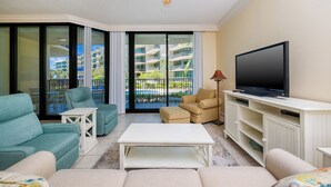 A living room with a flat-screen TV, a ceiling fan, and a balcony with a view of a building and palm trees. - Stay, Play, Make Memories Phoenix On The Bay 1123 (Orange Beach)