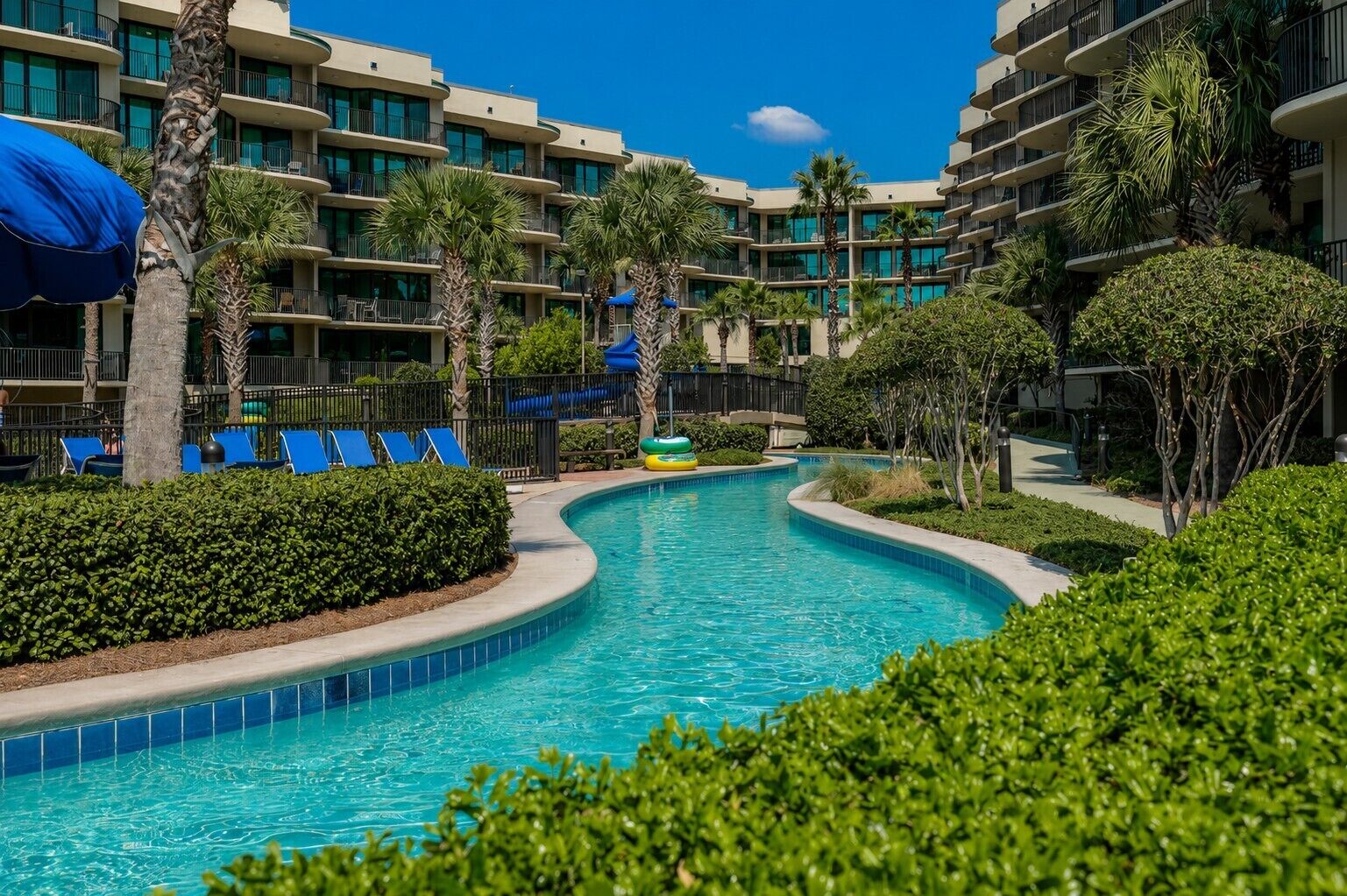 A hotel with a swimming pool, palm trees, and blue lounge chairs.