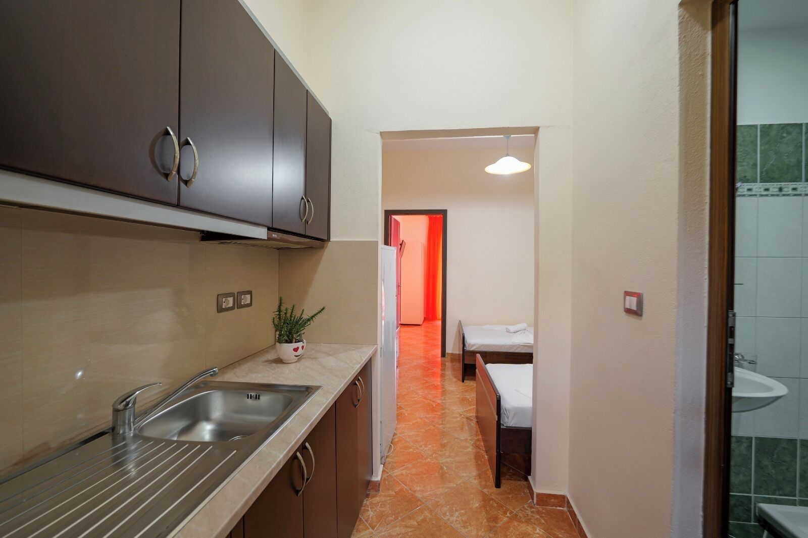 Un pasillo estrecho con una cocina a un lado, un comedor con mesa y sillas, y un baño con lavamanos e inodoro a la vista.