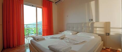 A hotel room with a large bed, a balcony with red curtains, and a view of the sea.