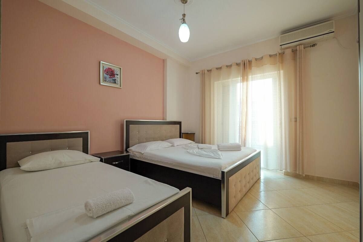 A hotel room with two beds, a small bedside table, a wall-mounted air conditioner, and a window with curtains.