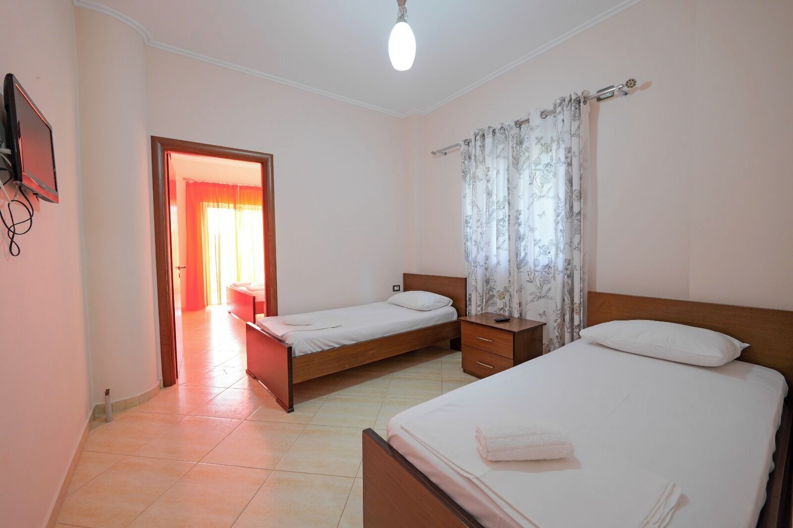 A hotel room with two single beds, a wooden nightstand, a small drawer, a TV mounted on the wall, and a bathroom visible through an open door.