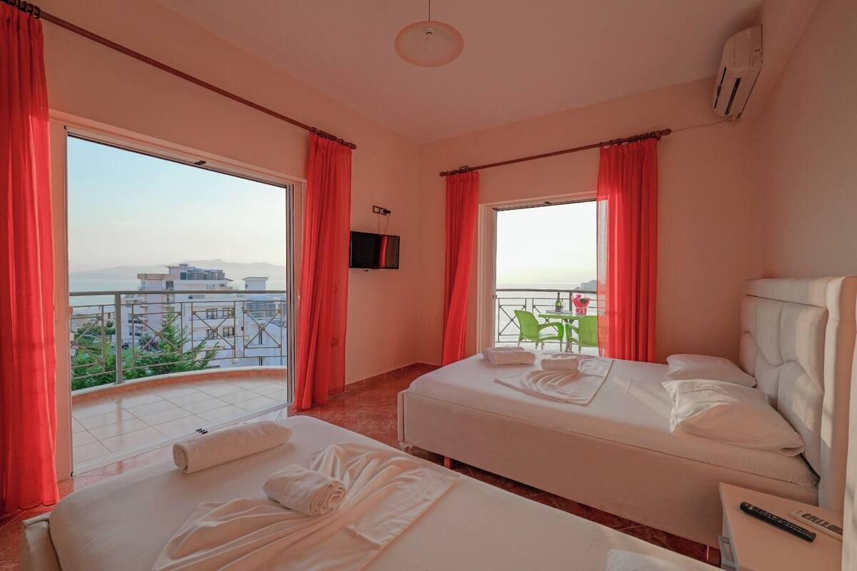 A hotel room with two beds, a balcony with red curtains, a TV, and a view of the city.