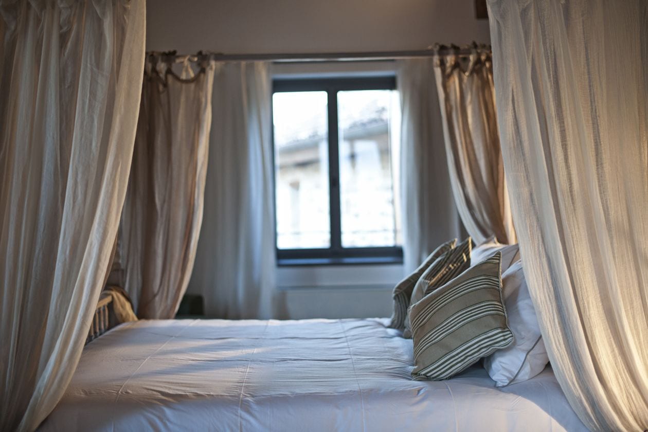 Ein Bett mit weißer Bettwäsche und gestreiften Kissen, ein Fenster mit Vorhängen und ein Blick auf Gebäude im Freien.