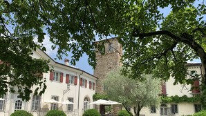 Une cour avec une tour en pierre, un bâtiment blanc aux volets rouges et des arbustes taillés avec soin.