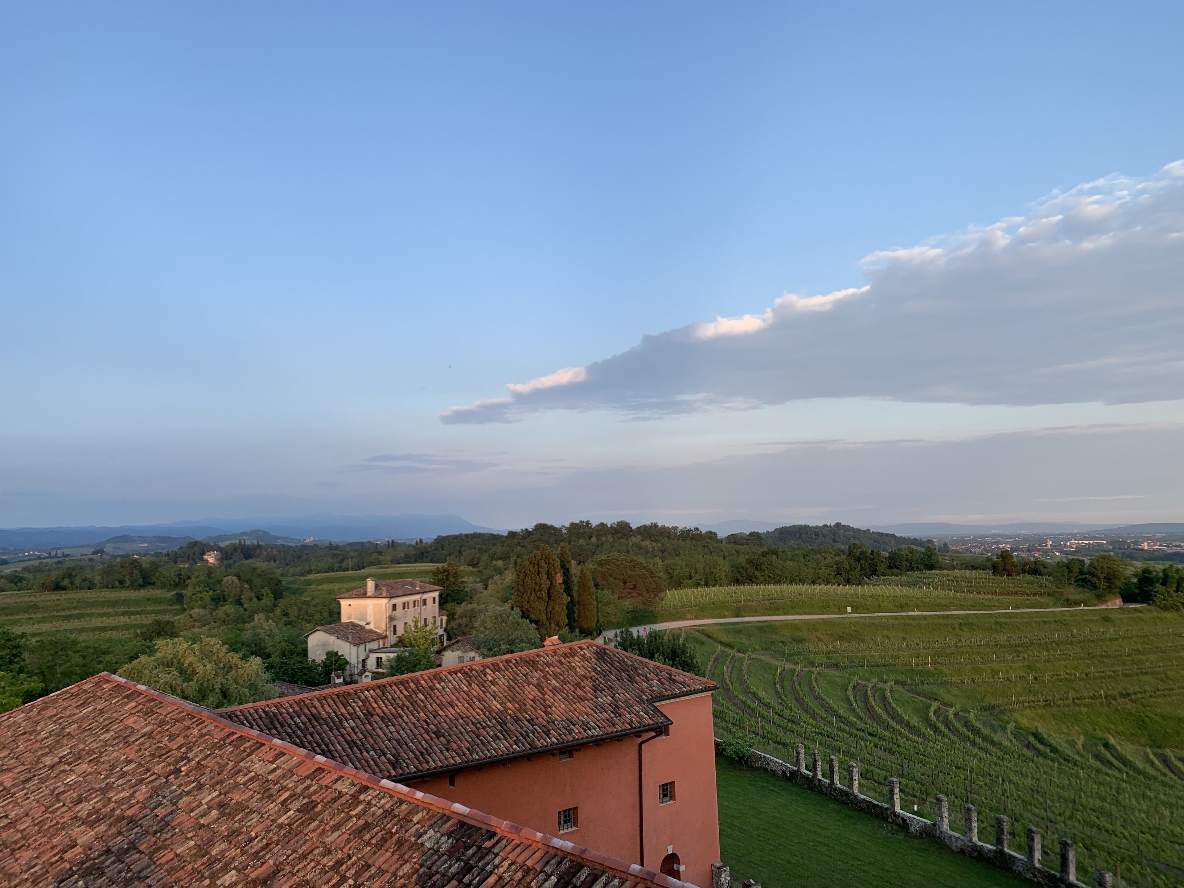 A rural landscape with terraced fields, a traditional building with a tiled roof, and a distant view of hills and a small town.