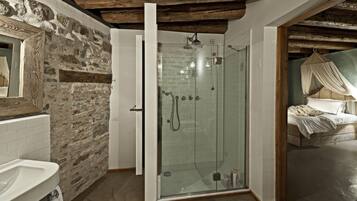 A rustic bathroom with a stone wall, a glass shower enclosure, and a bed with a canopy in the adjacent room.
