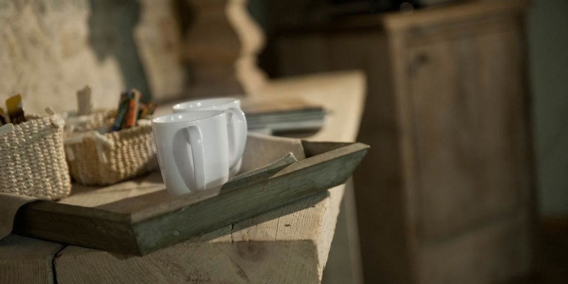 A wooden tray with two white mugs on a rustic table, a woven basket with pens, and a wooden cabinet in the background.