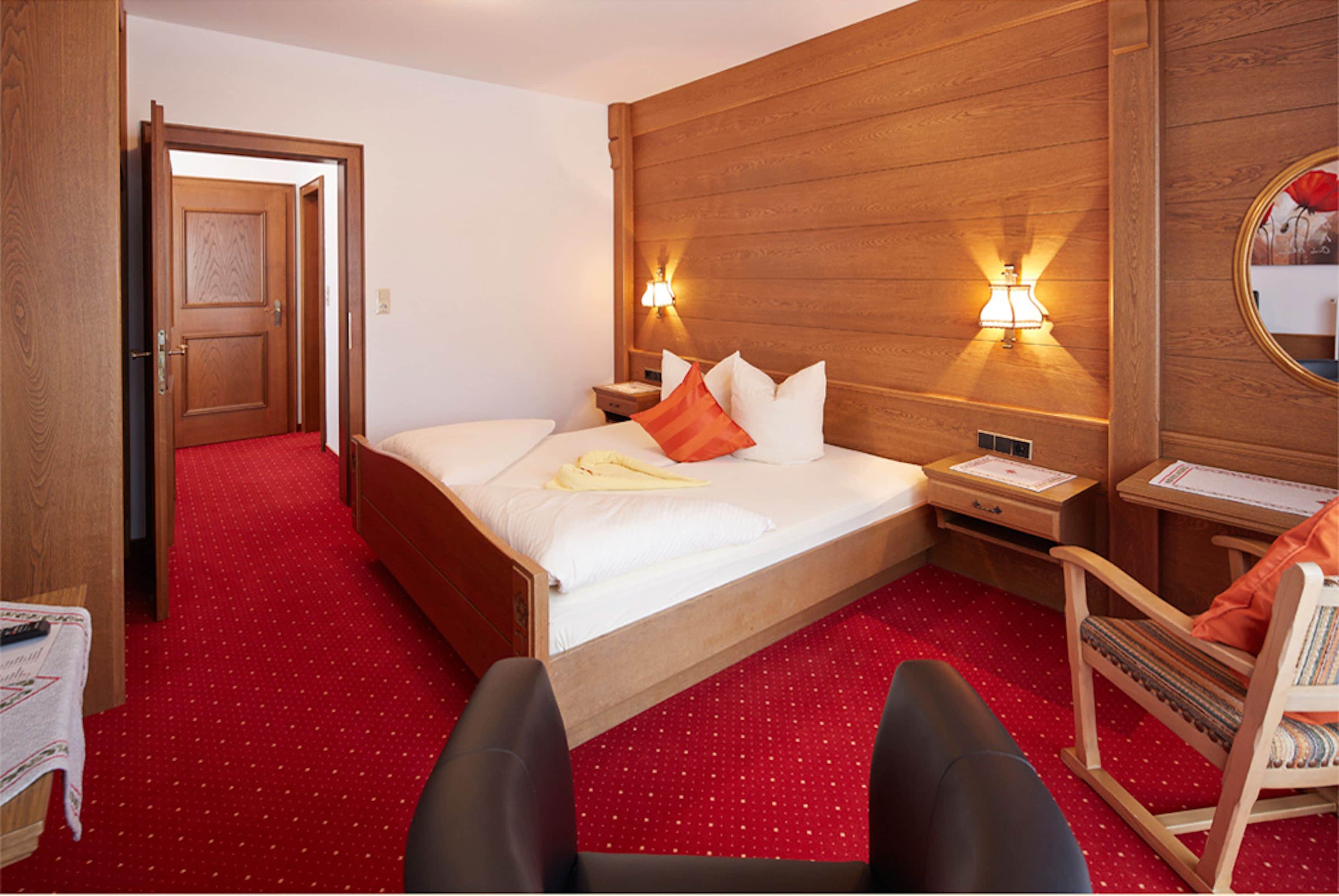 Une chambre d’hôtel avec une tête de lit en bois, un grand lit recouvert de draps blancs, deux tables de chevet avec des lampes, un fauteuil à bascule en bois et un sol recouvert d’une moquette rouge.