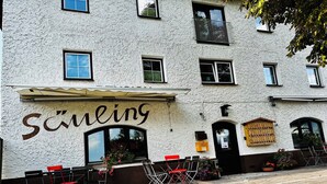Un edificio con el nombre 'Schüling' en la fachada, que presenta una hilera de sillas y mesas rojas en el exterior, un botellero de madera y un pequeño jardín con flores.