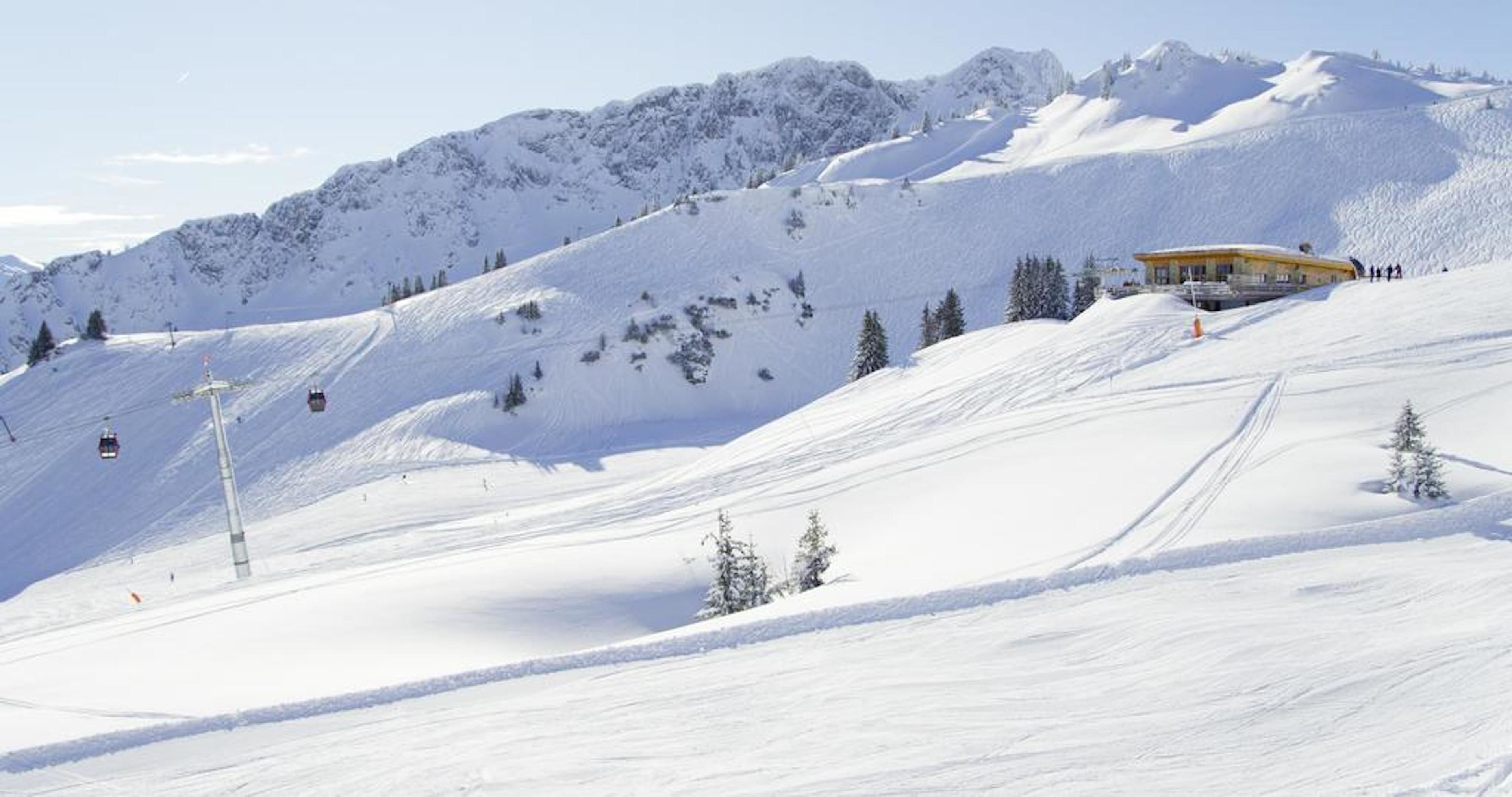 A snow-covered mountain slope with a ski lift, a yellow building, and a few skiers.