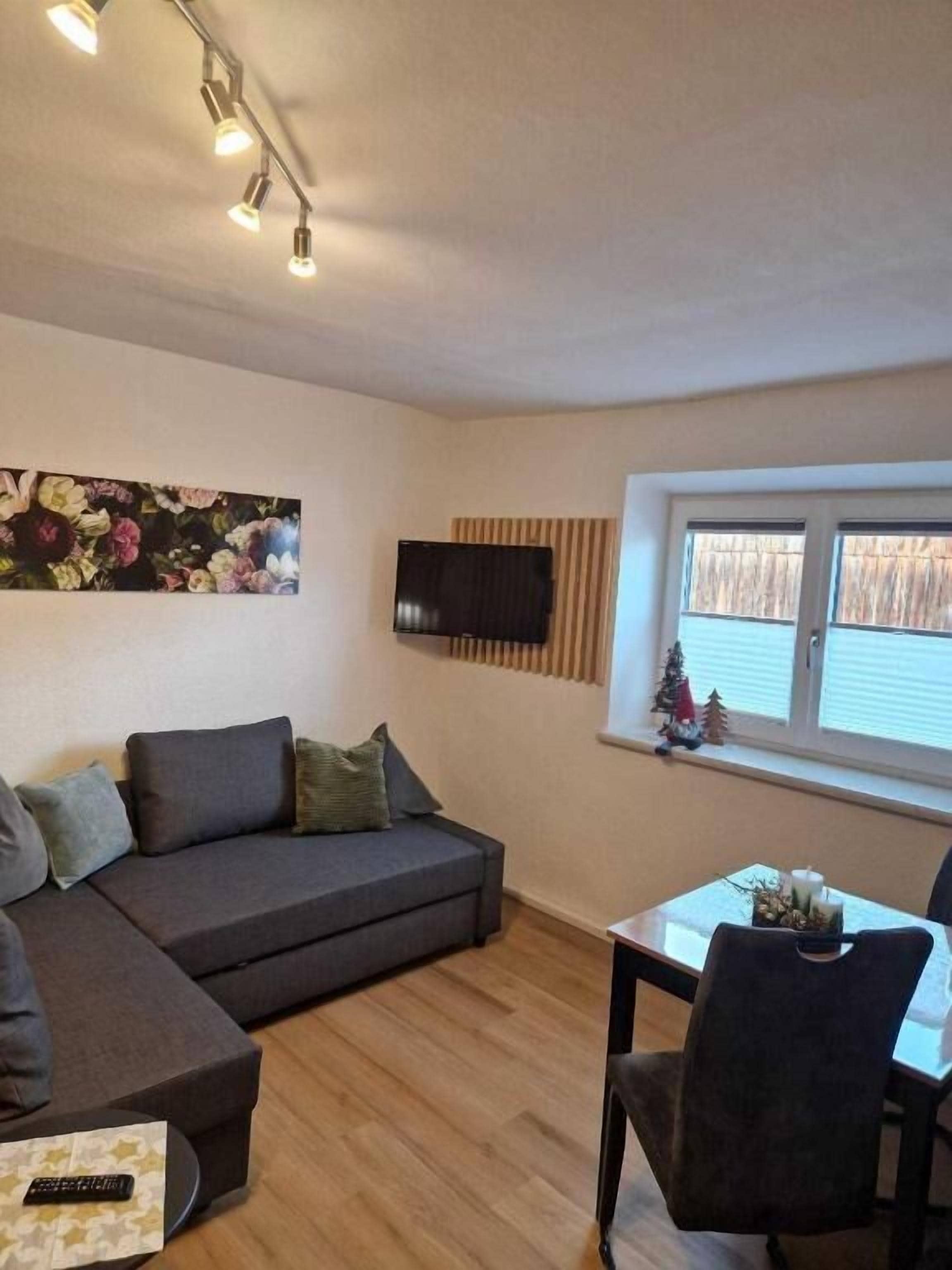 Una sala de estar acogedora con un sofá gris, una mesita con sillas, un televisor de pantalla plana montado en la pared y una ventana con persianas.