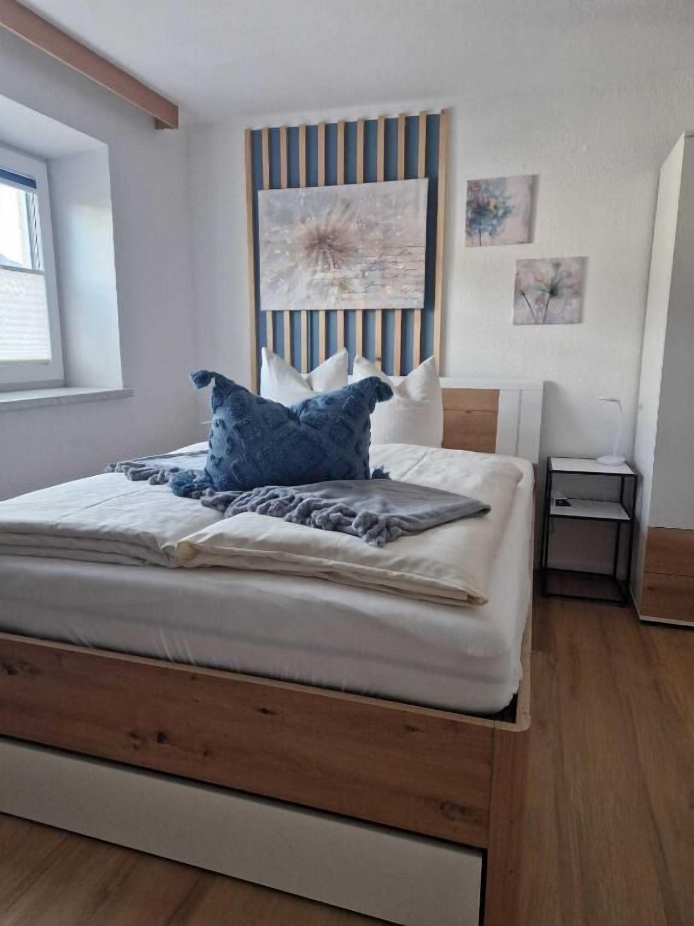 Una cama bien hecha con cabecera de madera, una mesita de noche con lámpara y una ventana con persianas.
