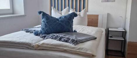 Una cama bien hecha con cabecera de madera, una mesita de noche con lámpara y una ventana con persianas.
