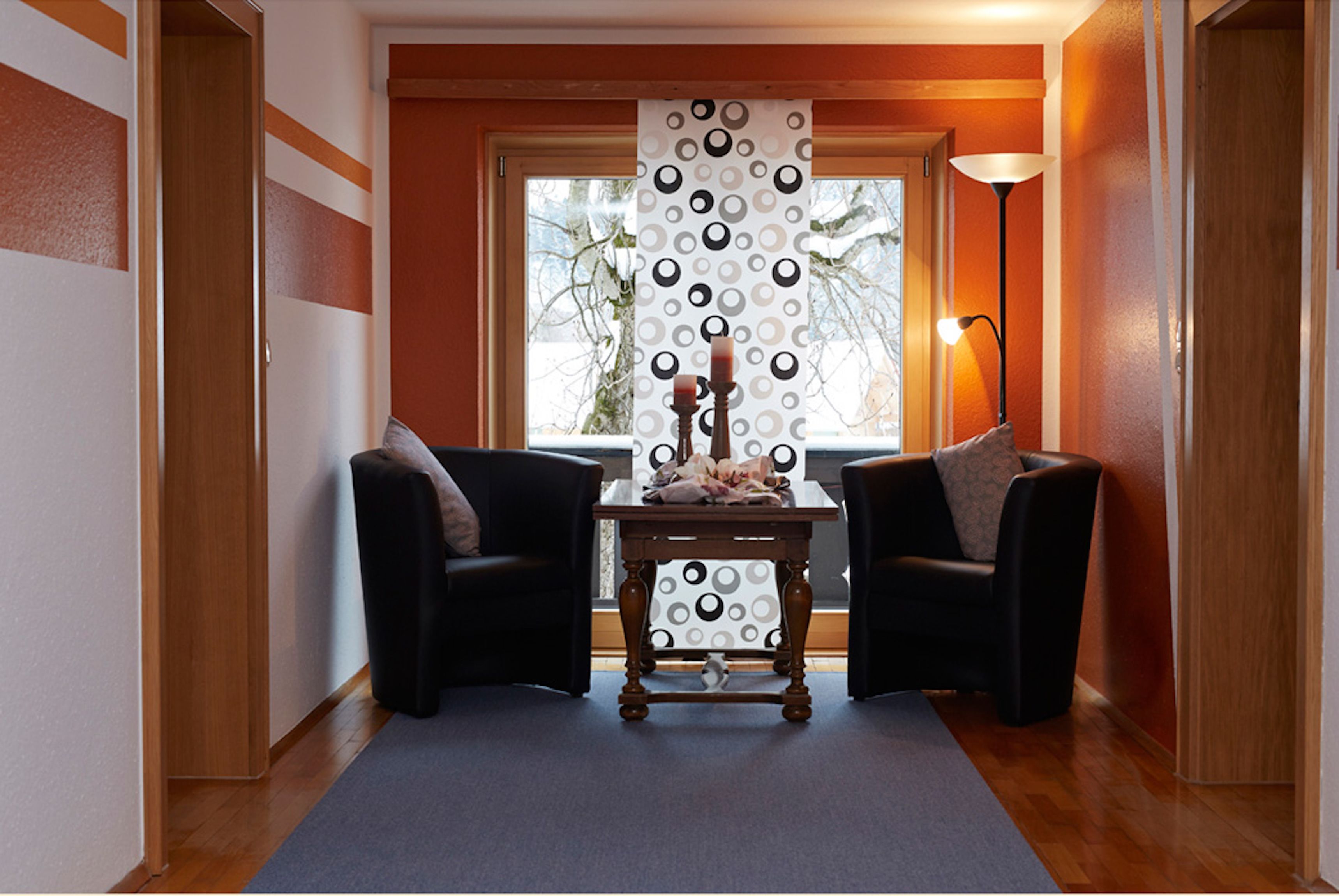 Una habitación con dos sillones negros, una mesa de madera con un florero y velas, una cortina estampada y una lámpara.