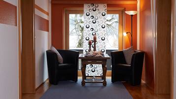 Una habitación con dos sillones negros, una mesa de madera con un florero y velas, una cortina estampada y una lámpara.