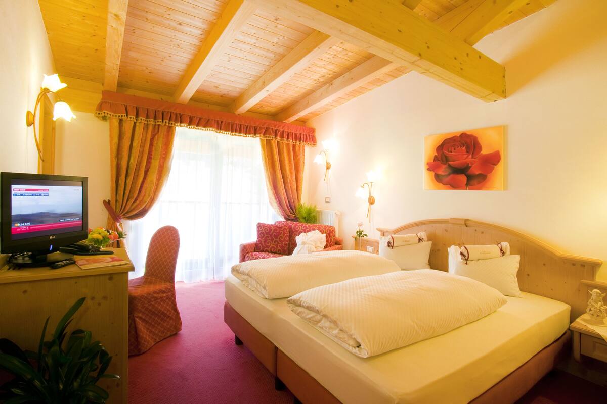 A hotel room with a wooden ceiling, a bed with white bedding, a desk with a computer, a chair, and a window with curtains.