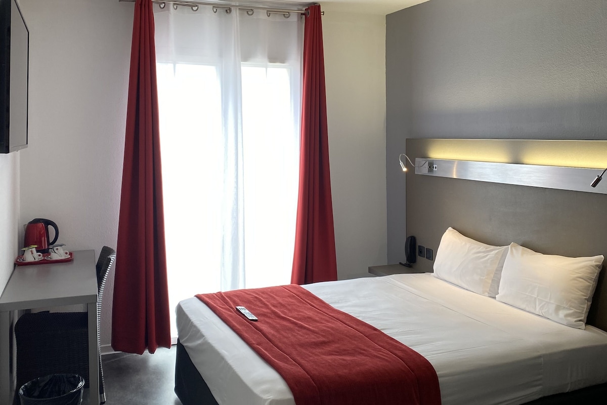 A hotel room with a bed, a desk, a chair, a trash bin, and a window with curtains.