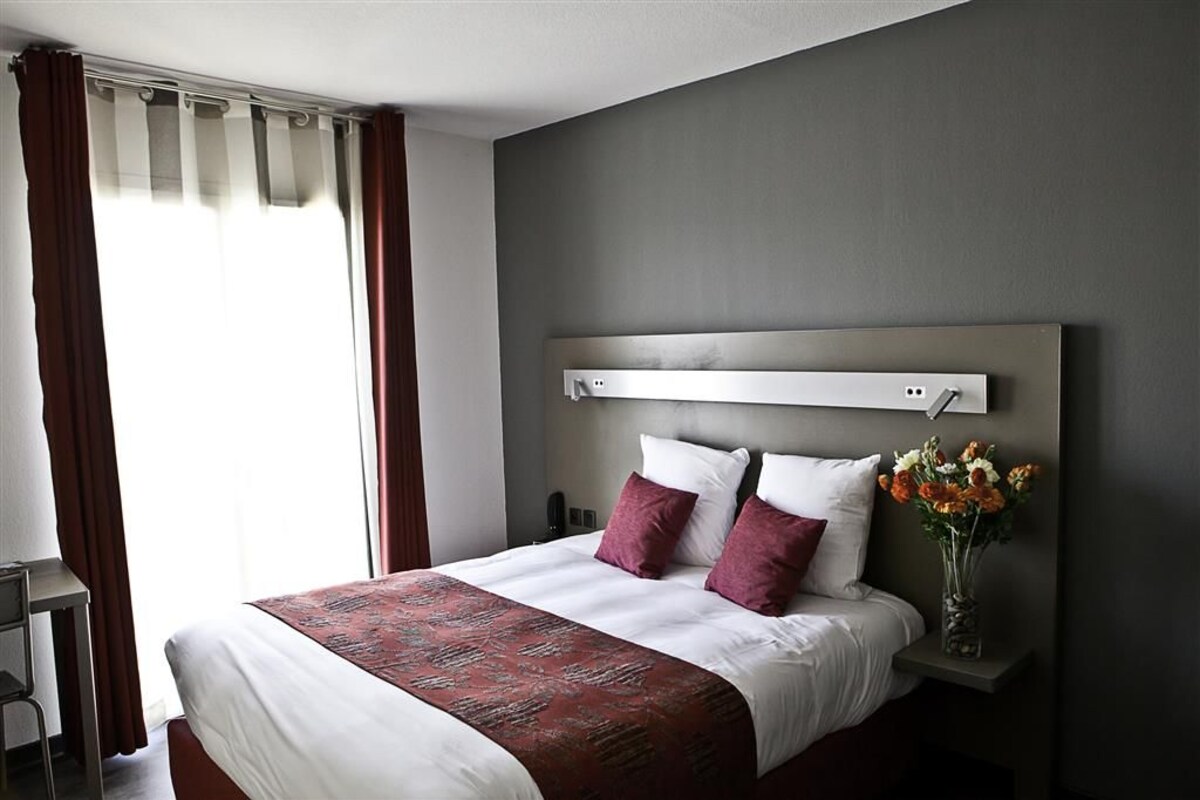 Habitació d'hotel amb un llit gran, una taula de nit i un taulell amb un ram de flors, i una finestra amb cortines.