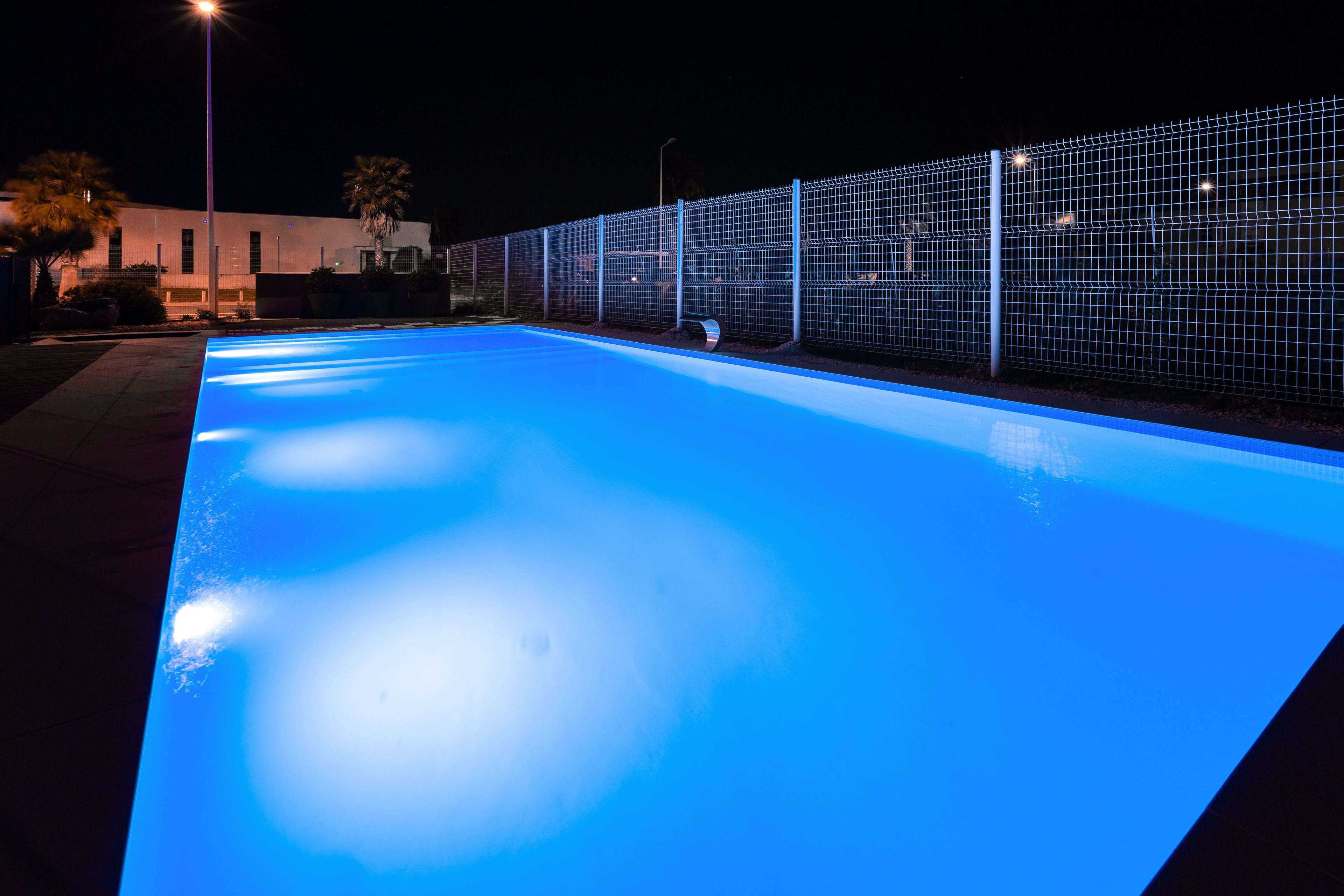 Una piscina con bordo illuminato di blu, circondata da una recinzione e un edificio sullo sfondo.