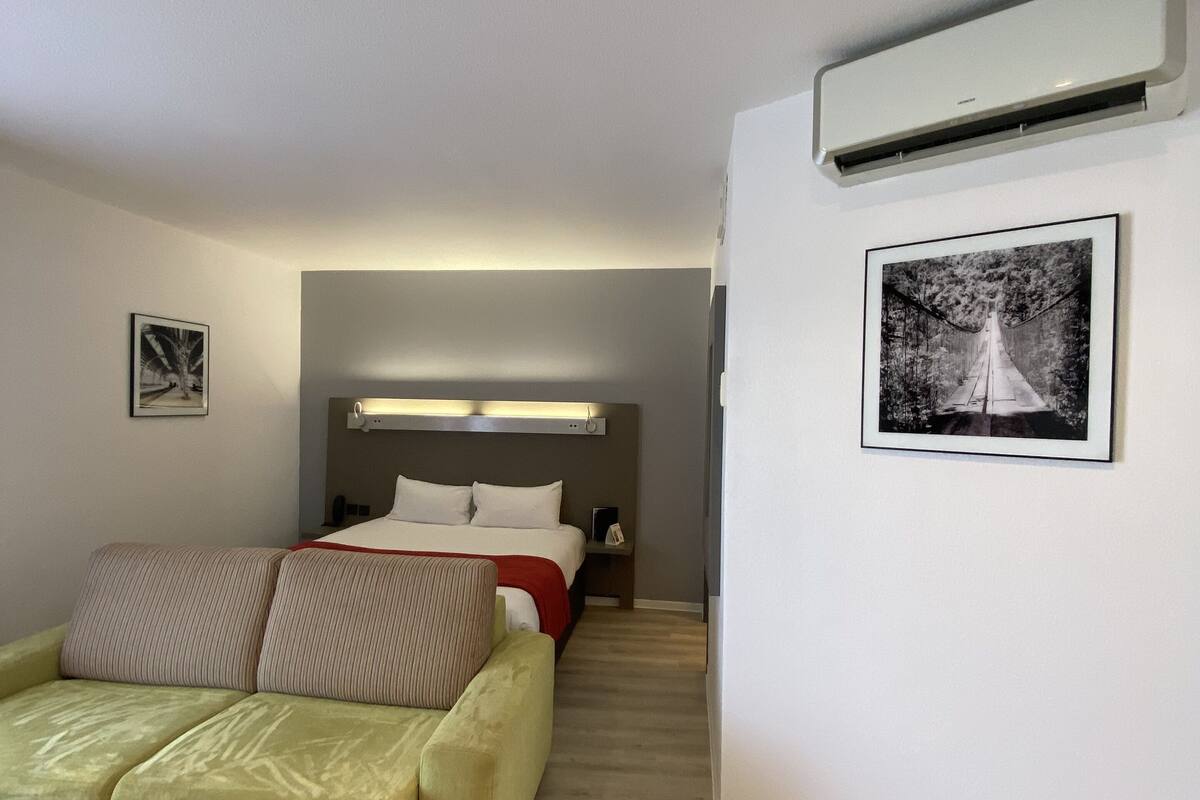 A modern hotel room with a bed, a sofa, a chair, and an air conditioning unit.