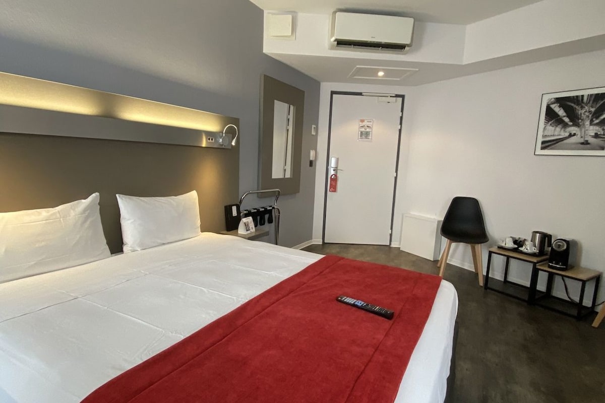 A hotel room with a large bed, a red throw blanket, a bedside table with a lamp, a chair, and a small table with a kettle and a speaker.