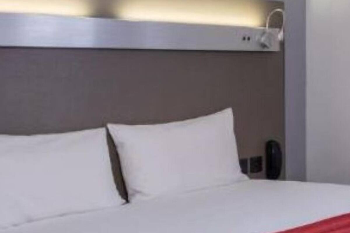 A modern hotel room with a neatly made bed, a red throw blanket, and a wall-mounted light fixture above the bed.