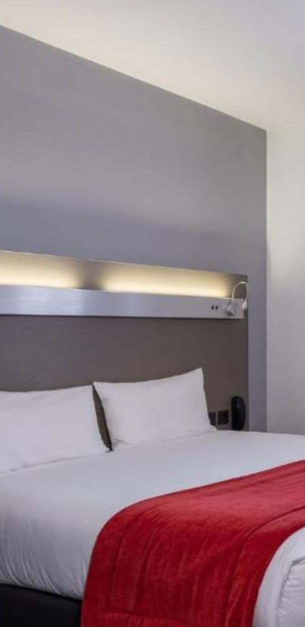 A hotel room with a bed, a red throw blanket, and a modern headboard with built-in lighting.