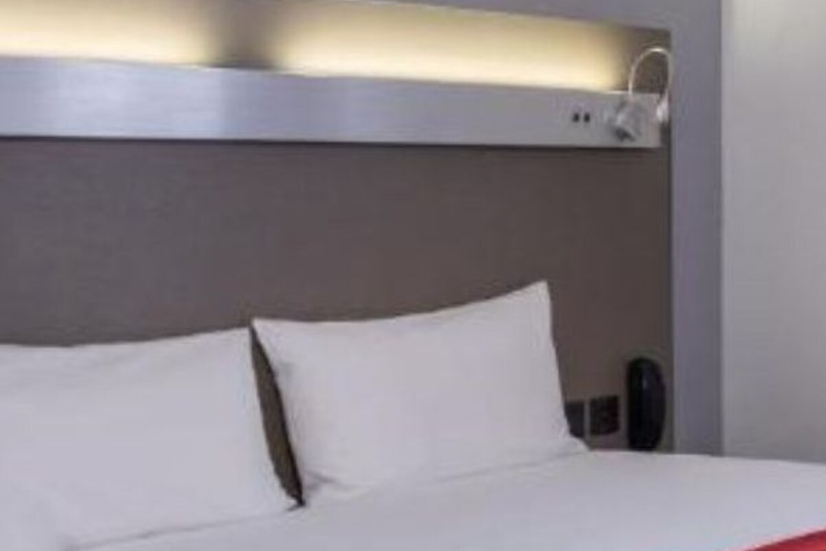 Una habitación de hotel moderna con una cama bien hecha, una manta roja y una lámpara de pared sobre la cama.