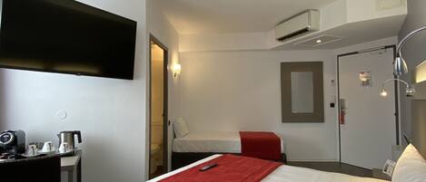 A hotel room with a bed, a desk, a chair, a TV, a mirror, and an air conditioning unit.