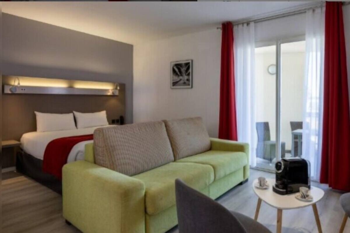 A hotel room with a bed, a green sofa, a coffee table with cups, and a sliding glass door with red curtains.