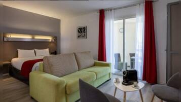 Une chambre d’hôtel avec un lit, un canapé vert, une table basse avec des tasses, et une porte-fenêtre coulissante avec des rideaux rouges.