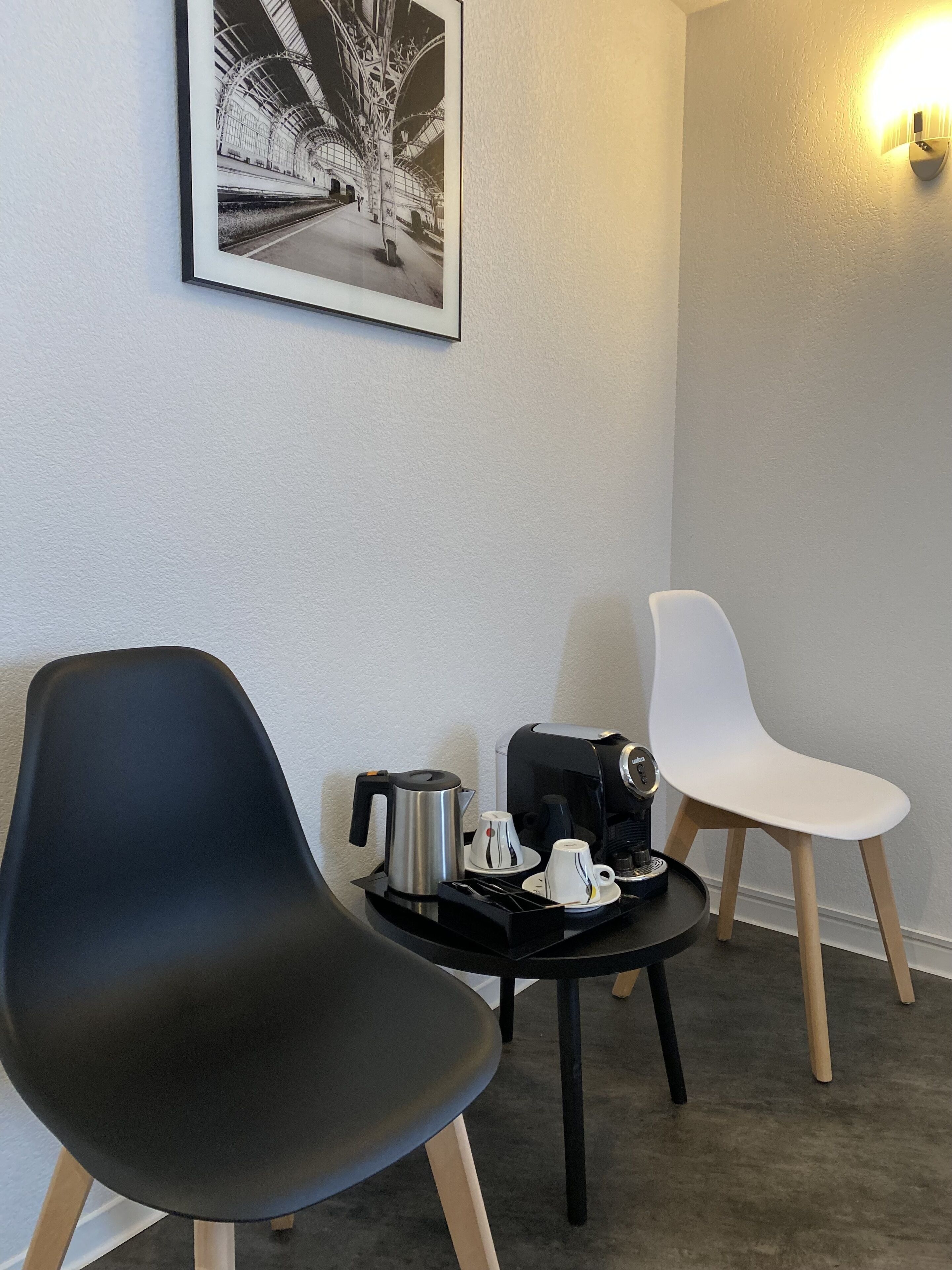 Een kamer met twee stoelen, een tafeltje met een koffiezetapparaat en kopjes, en een lijst aan de muur.
