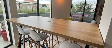 A modern dining area with a long wooden table, four white chairs, and large windows offering views of buildings and greenery.