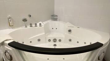 A large, round, white bathtub with multiple jets and a control panel, surrounded by white tiled walls and a brown floor.