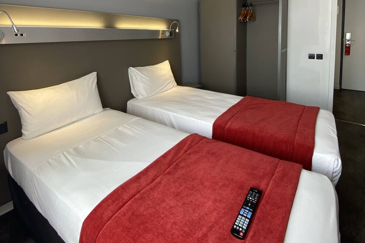 A hotel room with two beds, a bedside table, a wall-mounted lamp, and a wardrobe.