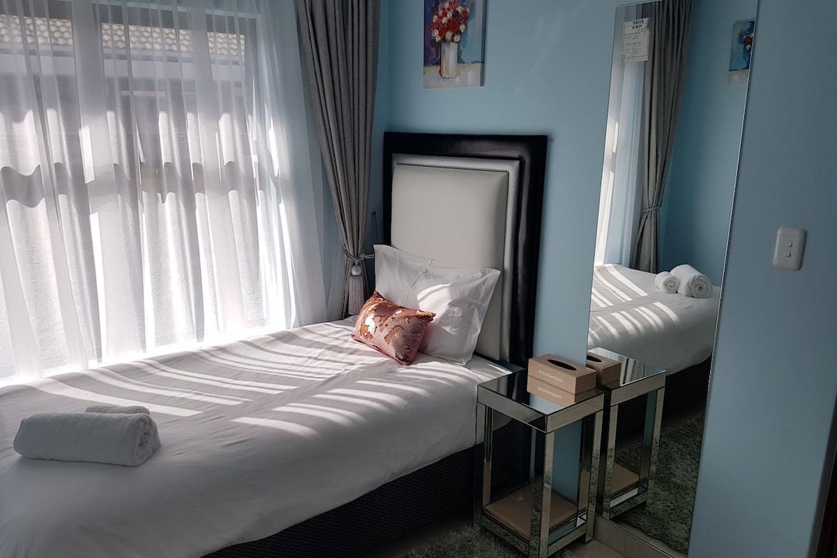 Ein ordentlich gemachtes Bett mit weißen Leinen und einem einzelnen Zierkissen, ein Nachttisch mit Taschentuchbox und ein Spiegel, der das Bett und die Vorhänge reflektiert.