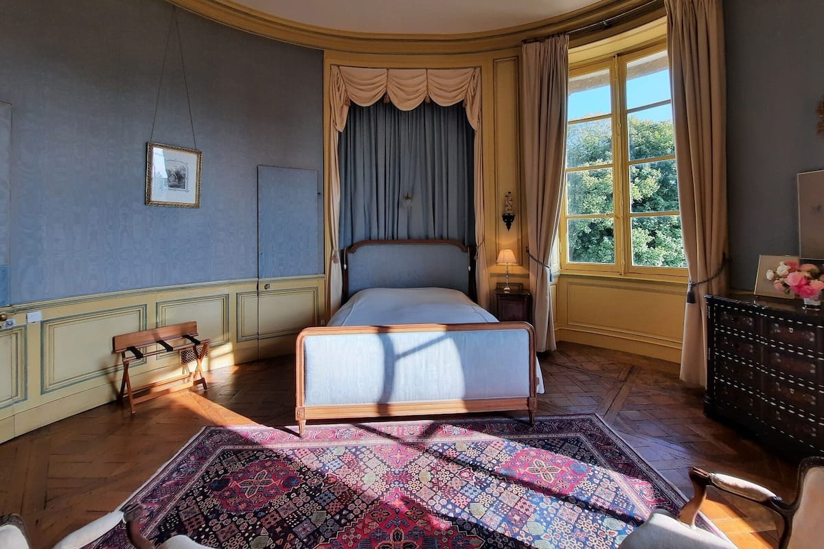 A bedroom with a bed, a nightstand, a dresser, a window with curtains, a patterned rug, and a chair.